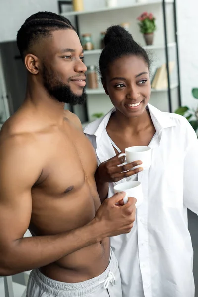 African american couple holding cups and looking away at kitchen — Stock Photo