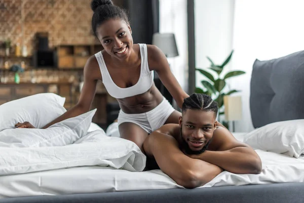 Улыбающаяся африканская американская пара в нижнем белье смотрит в камеру в спальне — стоковое фото