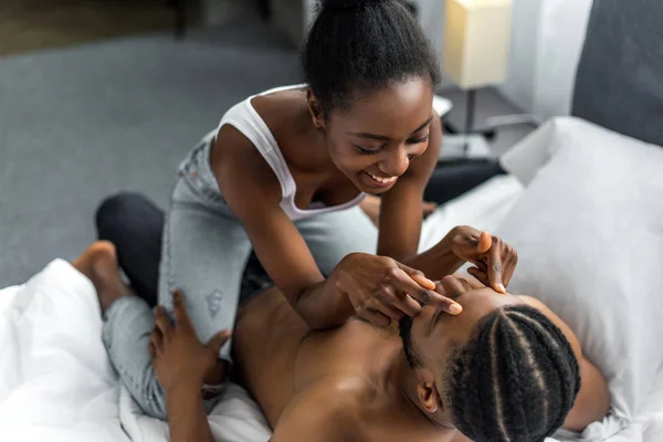 African american girlfriend sitting on boyfriend and touching forehead in bedroom — Stock Photo