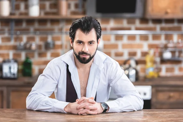 Guapo solitario hombre de negocios sentado en la mesa en la cocina y mirando a la cámara - foto de stock