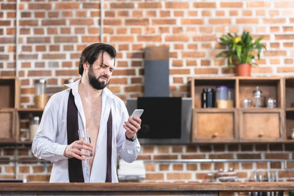 Guapo hombre de negocios solitario con resaca sosteniendo un vaso de agua y mirando el teléfono inteligente en la cocina - foto de stock