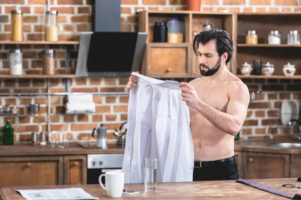Sin camisa guapo solitario hombre de negocios mirando camisa en la cocina - foto de stock