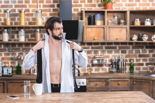 Guapo hombre de negocios solitario con camisa desabotonada sosteniendo corbata en la cocina - foto de stock