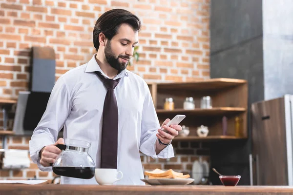 Guapo hombre de negocios solitario sosteniendo la olla con café y teléfono inteligente en la cocina — Stock Photo