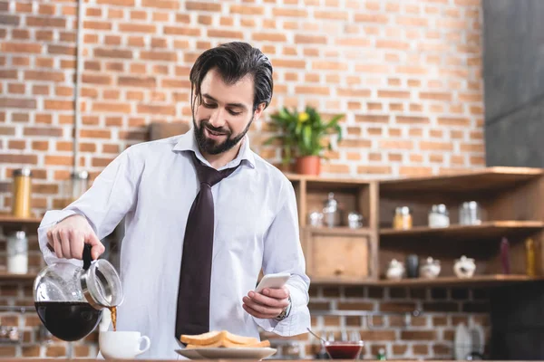 Красивый одинокий бизнесмен наливает кофе в чашку и держит смартфон на кухне — стоковое фото