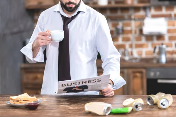 Обрезанный образ одинокого бизнесмена, пьющего кофе и читающего газету утром на кухне — стоковое фото