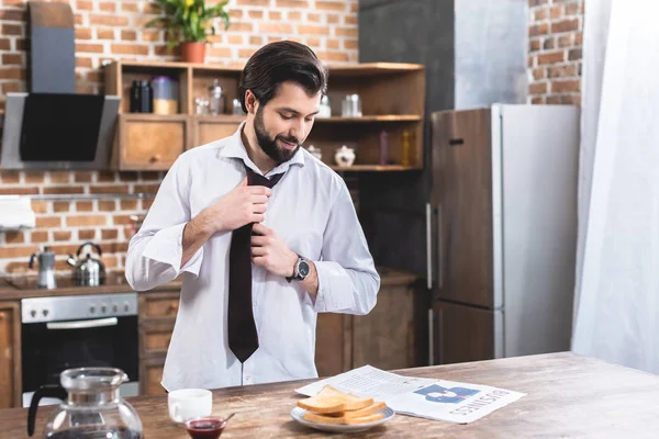 Handsome loner businessman reading newspaper and fixing tie at kitchen — Stock Photo