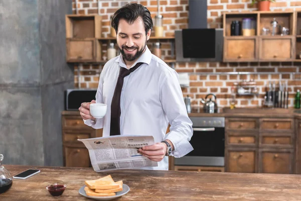 Guapo hombre de negocios solitario sosteniendo la taza de café y leyendo el periódico en la cocina - foto de stock