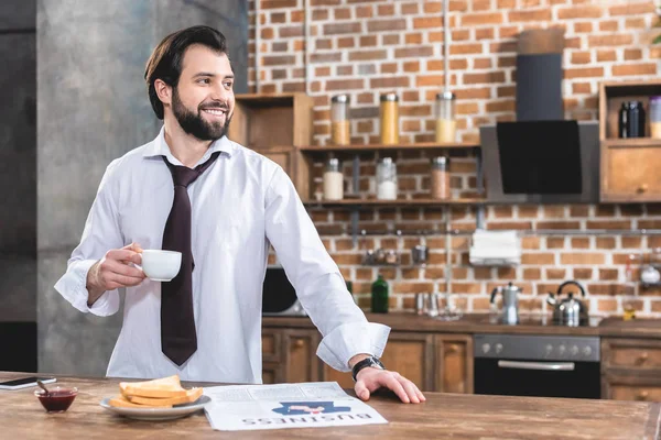 Handsome loner businessman holding cup of coffee and looking away at kitchen — Stock Photo
