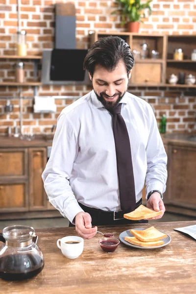 Bell'uomo d'affari solitario sorridente che aggiunge la marmellata su pane tostato a cucina — Foto stock