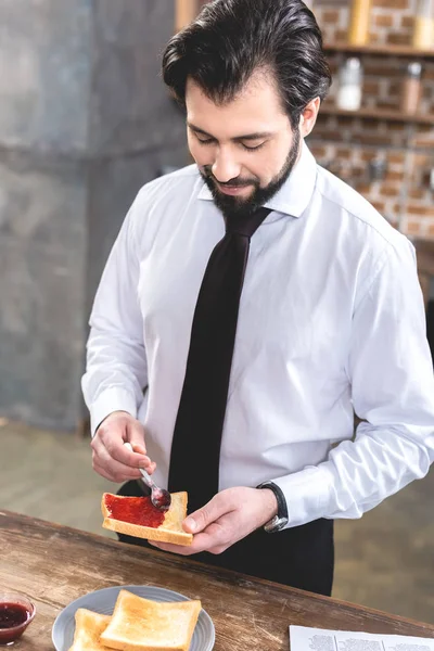 Bonito solitário empresário adicionando geléia em torradas na cozinha — Fotografia de Stock