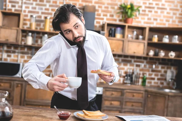 Loner businessman talking by smartphone while having breakfast at kitchen — Stock Photo