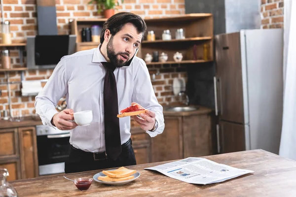 Guapo solitario hombre de negocios hablando por teléfono inteligente mientras desayuna en la cocina - foto de stock