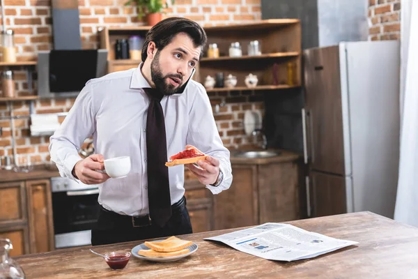 Bonito solitário empresário falando por smartphone enquanto tomando café da manhã na cozinha — Fotografia de Stock