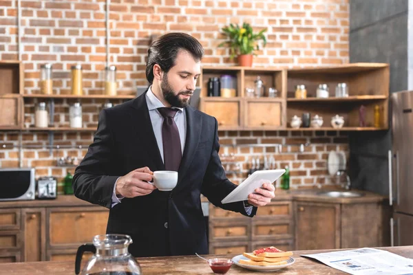 Guapo solitario hombre de negocios sosteniendo tableta y taza de café en la cocina - foto de stock