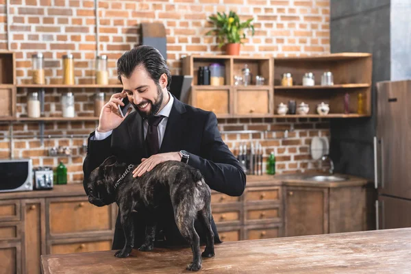 Handsome loner businessman talking by smartphone and palming dog at kitchen — Stock Photo