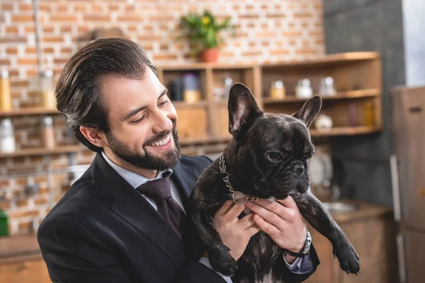 Sonriente guapo solitario hombre de negocios abrazando bulldog en cocina - foto de stock