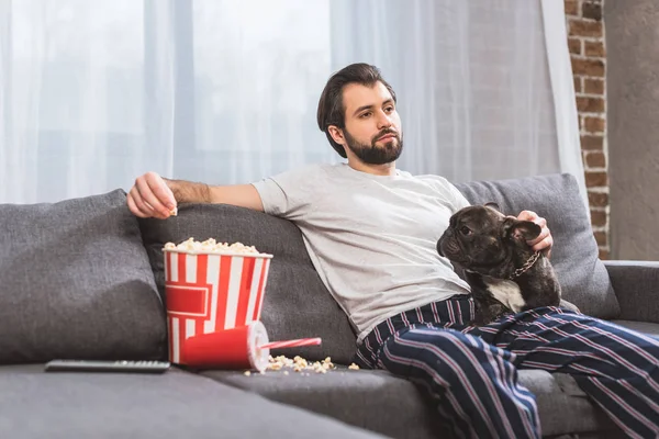 Handsome loner sitting with bulldog on sofa and eating popcorn in living room — Stock Photo