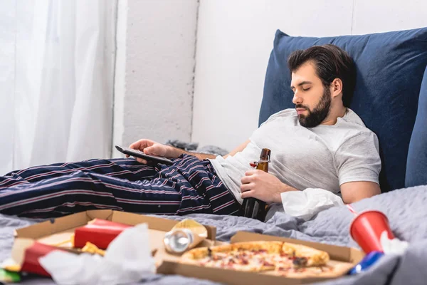 Handsome loner sleeping and holding remote control and bottle of beer in bedroom — Stock Photo