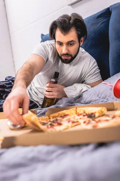 Guapo solitario tomando pedazo de pizza y sosteniendo botella de cerveza en la cama en el dormitorio - foto de stock