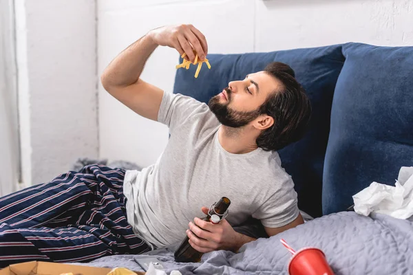 Schöner Einzelgänger, der Pommes frites isst und eine Flasche Bier im Schlafzimmer hält — Stockfoto