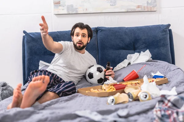 Schöner Einzelgänger gestikuliert, während er Fußballspiel mit Bier im Schlafzimmer anschaut — Stockfoto