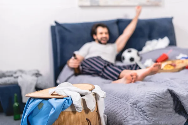 Bello solitario guardando partita di calcio con birra in camera da letto con cesto di lavanderia in primo piano — Foto stock