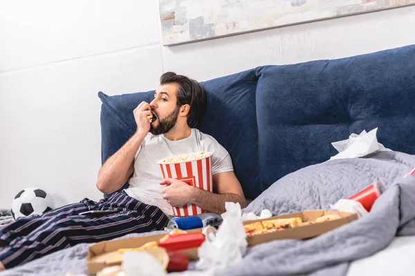 Guapo solitario comiendo palomitas de maíz y viendo la televisión en el dormitorio - foto de stock
