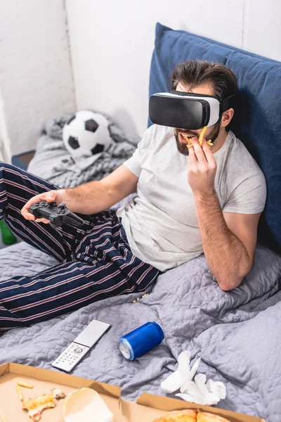 Solitário usando fone de ouvido realidade virtual, jogando videogame e comendo batatas fritas no quarto — Fotografia de Stock