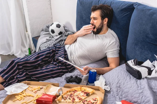 Handsome loner holding tablet and yawning on bed in bedroom — Stock Photo