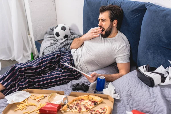 Schöner Einzelgänger mit Tablette und Gähnen auf dem Bett im Schlafzimmer — Stockfoto