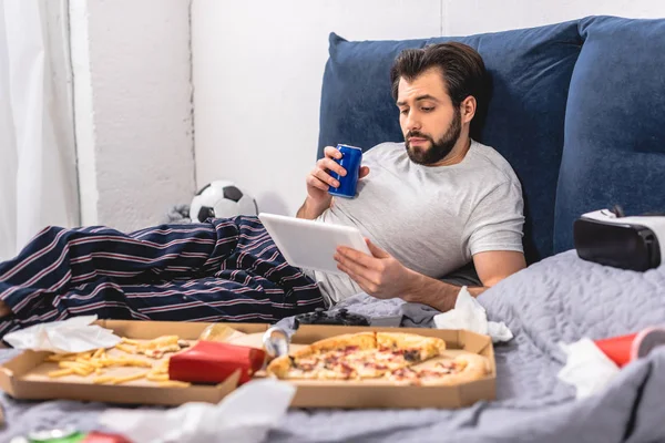 Beau solitaire utilisant la tablette et tenant boisson sur le lit dans la chambre — Photo de stock