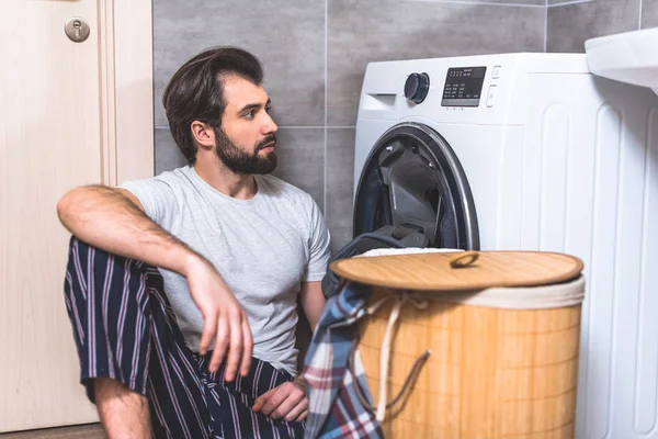 Bonito solitário sentado no chão e olhando para máquina de lavar roupa no banheiro — Fotografia de Stock