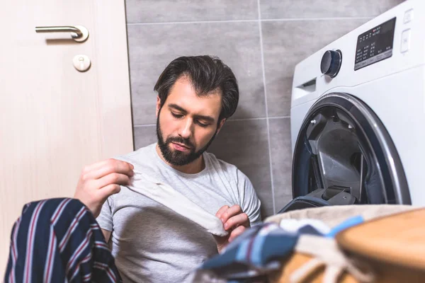 Guapo solitario sosteniendo calcetín sucio cerca de la lavadora en el baño - foto de stock