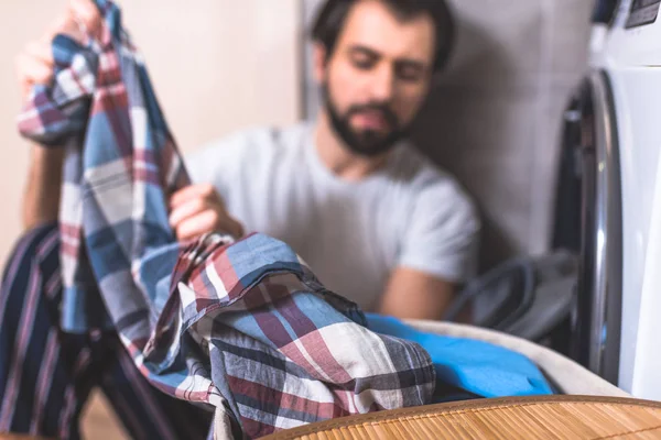 Красивий учень бере одяг для прання біля пральної машинки у ванній кімнаті з сорочкою на передньому плані — стокове фото