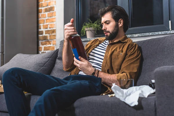 Bello solitario guardando bottiglia di bevanda alcolica in soggiorno — Foto stock