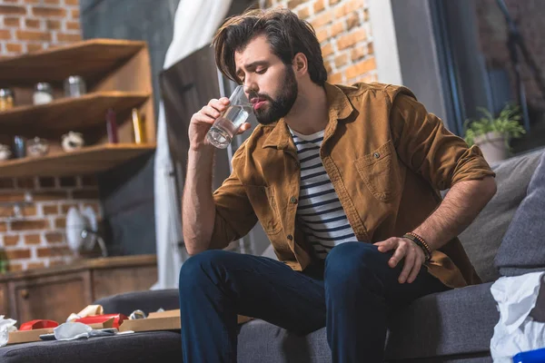 Schöner Einzelgänger trinkt Wasser, während er morgens im Wohnzimmer Kater hat — Stockfoto