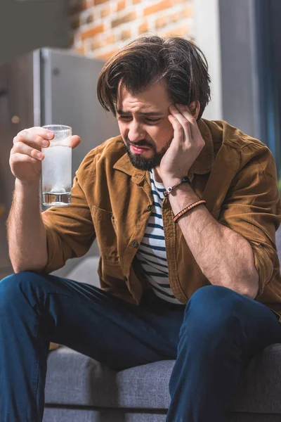 Schöner Einzelgänger, der ein Glas Wasser mit Tablette in der Hand hält und Kopfschmerzen im Wohnzimmer hat — Stockfoto