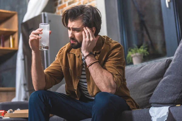 Solitaria con dolor de cabeza y resaca y mirando un vaso de agua con pastillas en la sala de estar - foto de stock