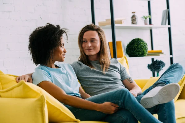 Sorrindo casal multicultural olhando um para o outro e sentado no sofá — Fotografia de Stock