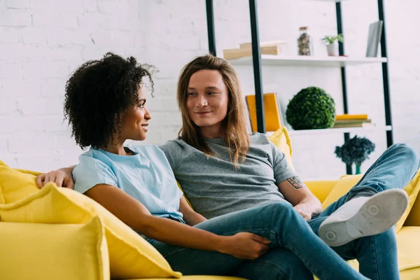 Couple multiculturel souriant se regardant et assis sur le canapé — Photo de stock