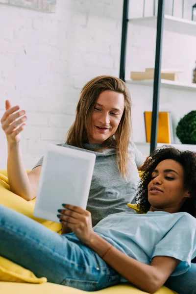 Sorrindo jovem casal multiétnico com tablet digital no sofá — Fotografia de Stock