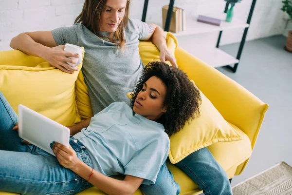 Молодая межрасовая пара на диване с цифровым планшетом и чашкой кофе — стоковое фото
