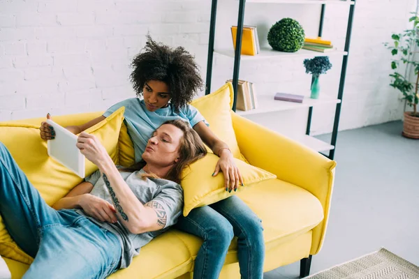 Young multicultural couple on couch looking at digital tablet screen — Stock Photo