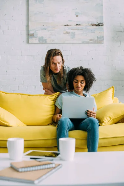 Attraente donna afro-americana utilizzando il computer portatile sul divano e fidanzato in piedi dietro — Foto stock