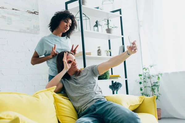 Junger Mann gestikuliert und macht Selfie auf Smartphone mit afrikanisch-amerikanischer Freundin, die Friedenszeichen macht — Stockfoto