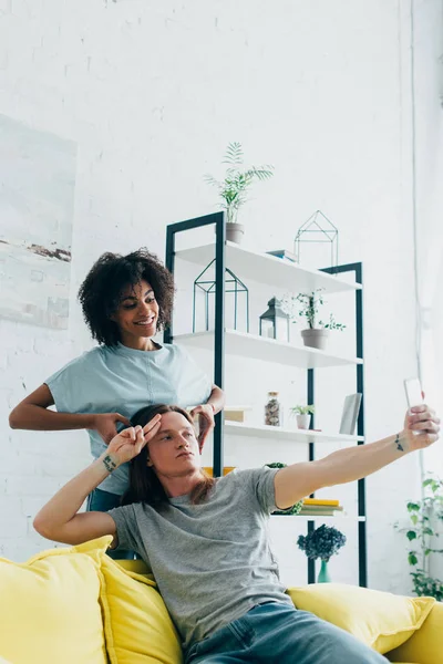 Multiculturel jeune couple geste et prendre selfie sur smartphone à la maison — Photo de stock