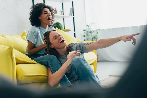 Riéndose joven con taza de café apuntando en la pantalla de televisión a la novia afroamericana sonriente - foto de stock