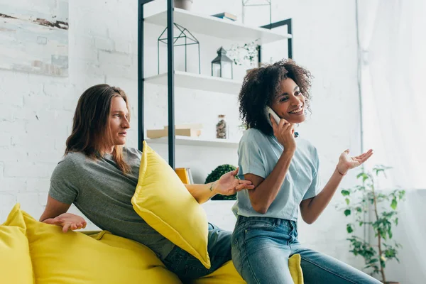 Sonriente mujer afroamericana hablando en smartphone mientras su novio la mira con los brazos anchos - foto de stock