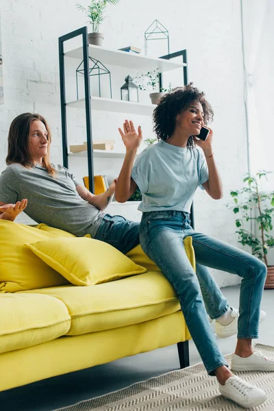 Sonriente mujer afroamericana hablando en smartphone y haciendo gestos a mano a novio ofendido - foto de stock